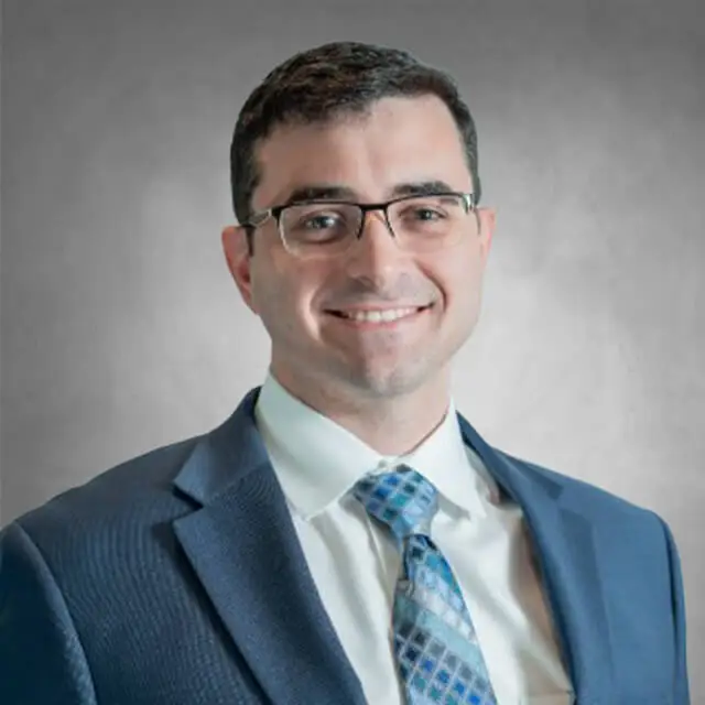 Headshot of a man wearing glasses, a dark suit jacket, light blue shirt, and a blue plaid tie against a gray studio background.